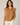 A woman wearing a tan short-sleeve blouse with a split neckline and pleated details, paired with off-white pants, against a neutral beige background.
