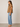 A woman stands in profile wearing a sleeveless tan top, light blue wide-leg jeans, and tan heeled sandals against a plain light beige studio wall and floor.