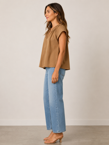 A woman stands in profile wearing a sleeveless tan top, light blue wide-leg jeans, and tan heeled sandals against a plain light beige studio wall and floor.