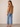 A woman modeling a brown sleeveless top with ruffled trim at the neckline, paired with light blue cropped jeans and beige heeled sandals, shown from a side-back angle against a plain beige studio background.