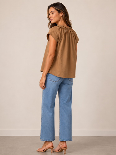 A woman modeling a brown sleeveless top with ruffled trim at the neckline, paired with light blue cropped jeans and beige heeled sandals, shown from a side-back angle against a plain beige studio background.