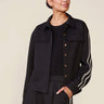 A woman modeling a black cropped button-front jacket with white double stripes down the sleeves, paired with matching black pants, against a plain beige studio background.