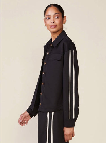 A woman modeling a black cropped button-front jacket with white double stripes down the sleeves, paired with matching black pants, against a plain beige studio background.