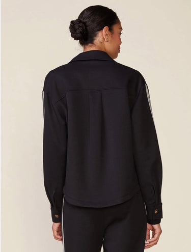 A woman shown from the back wearing a black long-sleeve collared jacket with subtle sheer side panels and black pants, photographed against a plain light beige studio background.
