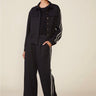 A model wearing a black two-piece tracksuit with white double stripes down the sleeves and legs, featuring a cropped button-front jacket and matching wide-leg pants, paired with white sneakers against a solid beige background.