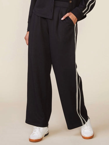 Black wide-leg track pants with double white stripes down the side, modeled with white sneakers against a light beige background.