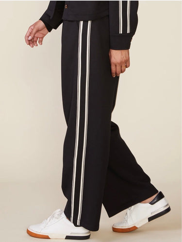 Black wide-leg sweatpants with double white side stripes, shown on a model from the side wearing white sneakers against a beige background.