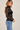 A woman wearing a long sleeve lace sheer top with a black bandeau and blue jeans on a neutral background.