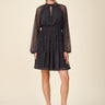 A woman modeling a short black mesh dress with a white polka dot pattern, long sheer sleeves, and a high keyhole neckline, paired with black slingback heels against a solid beige background.