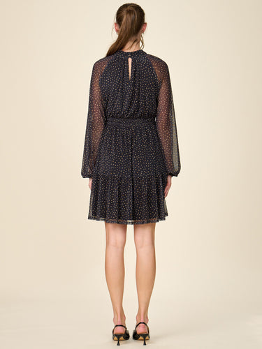 A model viewed from the back wearing a sheer black long-sleeve mini dress with a small white polka dot pattern, a keyhole back, and a tiered skirt, paired with black high heels against a solid beige background.