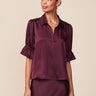 A woman modeling a dark plum satin blouse with short ruffled sleeves and a split V-neckline, paired with a matching satin skirt against a solid beige background.