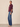 A woman stands in profile wearing a dark plum button-up blouse with ruffled elbow-length sleeves and blue wide-leg jeans against a plain beige studio background.