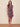 A woman modeling a fitted knee-length dress with a floral pattern in dusty purple, mauve, and pink, holding a small bright pink clutch against a beige studio backdrop.