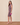 A woman models a sleeveless knee-length dress with a purple and pink floral pattern, paired with nude heels and a small pink clutch, standing against a beige studio backdrop.