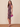 A woman modeling a sleeveless ruched knee-length dress with a dark purple and pink floral pattern, holding a bright pink clutch against a beige studio backdrop.