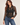 A woman modeling a sheer black long-sleeve button-up blouse with intricate floral lace and bead applique on the front, paired with blue jeans against a light beige studio background.