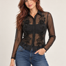A woman modeling a sheer black long-sleeve button-up blouse with intricate floral lace and bead applique on the front, paired with blue jeans against a light beige studio background.