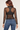 A woman shown from the back wearing a sheer black long-sleeve mesh top over a black sports bra, paired with blue jeans, against a light beige studio background.