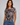 A woman modeling a short-sleeve crewneck t-shirt with a muted geometric block pattern in gray, brown, and rust tones, paired with blue jeans against a solid light gray background.