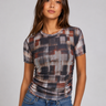 A woman modeling a short-sleeve crewneck t-shirt with a muted geometric block pattern in gray, brown, and rust tones, paired with blue jeans against a solid light gray background.