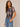 A woman modeling a short-sleeve crewneck top with a muted geometric plaid pattern in gray, brown, and burgundy, paired with light blue jeans against a neutral beige background.