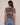 A woman viewed from the back wearing a short-sleeve ruched top with an abstract pixelated pattern in brown, gray, and black, paired with blue jeans against a plain beige background.