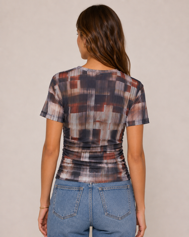 A woman viewed from the back wearing a short-sleeve ruched top with an abstract pixelated pattern in brown, gray, and black, paired with blue jeans against a plain beige background.