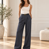 A woman stands in a bright, modern room wearing high-waisted, wide-leg navy pinstripe trousers with a gold-tone belt, paired with a white tank top and black loafers.