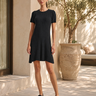A woman modeling a short-sleeve black ribbed knit dress with a flared skirt, paired with black loafers, standing on a sunlit patio with a large planter and stone wall in the background.