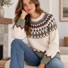 A woman sits indoors wearing a cream knit sweater with a Fair Isle style pattern in olive green, dark brown, and burgundy, paired with blue jeans.