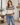 A woman wearing a cream knit sweater with a brown and burgundy geometric pattern across the chest and green ribbed cuffs and hem, paired with blue jeans in a bright living room.