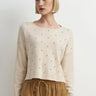 A woman wearing an embellished cream knit sweater and mustard yellow pants on a white background.