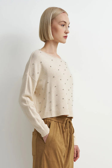 A woman wearing an embellished cream knit sweater and mustard yellow pants on a white background.