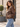 A woman wearing a brown faux-leather jacket with a tan sherpa collar and cuffs, paired with light blue jeans, standing outdoors in front of a beige building.