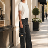 A woman stands on a sunlit city sidewalk wearing a white short-sleeve knit top, dark wide-leg embellished jeans, black shoes, and holding a small black handbag.