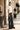 A woman stands on a sunlit city sidewalk wearing a white short-sleeve knit top, dark wide-leg embellished jeans, black shoes, and holding a small black handbag.