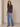 A woman modeling a dark brown long-sleeve button-front cardigan with light-wash blue jeans and brown pointed-toe heels, standing against a neutral beige studio wall.