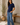 A woman modeling a navy blue short-sleeve ribbed knit top and wide-leg blue jeans, standing in a sunlit room with a potted plant and a wooden bench in the background.