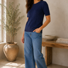 A woman modeling a navy blue short-sleeve ribbed knit top and wide-leg blue jeans, standing in a sunlit room with a potted plant and a wooden bench in the background.