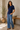 A woman modeling a navy blue short-sleeve ribbed knit top and wide-leg blue jeans, standing in a sunlit room with a potted plant and a wooden bench in the background.