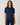 A woman wearing a navy blue short-sleeve cable-knit sweater and dark blue jeans, standing against a light gray studio background.