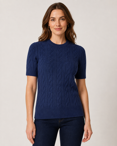 A woman wearing a navy blue short-sleeve cable-knit sweater and dark blue jeans, standing against a light gray studio background.