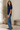 A woman wearing a navy blue short-sleeve cable-knit top and light blue wide-leg jeans, standing in a bright, neutral-toned room with a potted plant and a wooden bench.