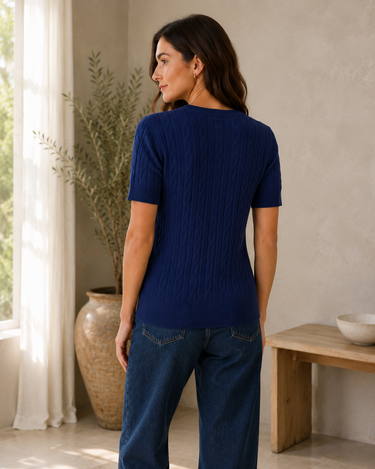 A woman wearing a short-sleeve navy blue ribbed knit top and dark blue jeans, photographed from the back in a bright, modern room with a window, potted plant, and wooden bench.