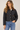 A woman wearing a black diamond-quilted jacket with a brown collar and pocket trim, paired with blue jeans, against a light beige studio background.