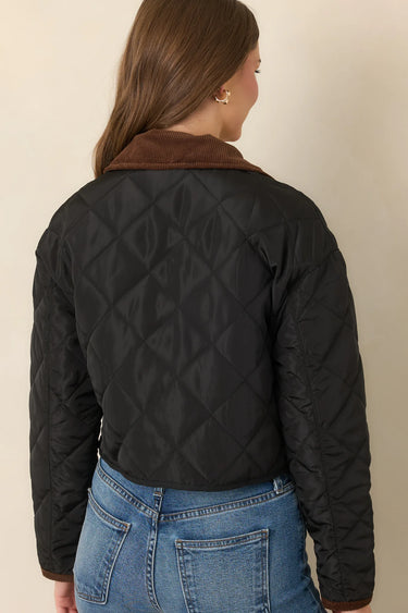 a woman wearing a black quilted jacket with a brown corduroy collar on a neutral background.
