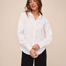 A woman wearing a white long-sleeve button-up shirt and black pants, standing against a solid beige studio background.