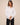 A woman wearing a white long-sleeve button-up shirt and black pants, standing against a solid beige studio background.