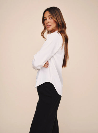 A woman posing in a white long-sleeve blouse and black wide-leg trousers against a solid beige studio background.