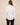 A woman viewed from the back wearing a white long-sleeve button-up shirt and black pants, standing against a plain beige studio background.